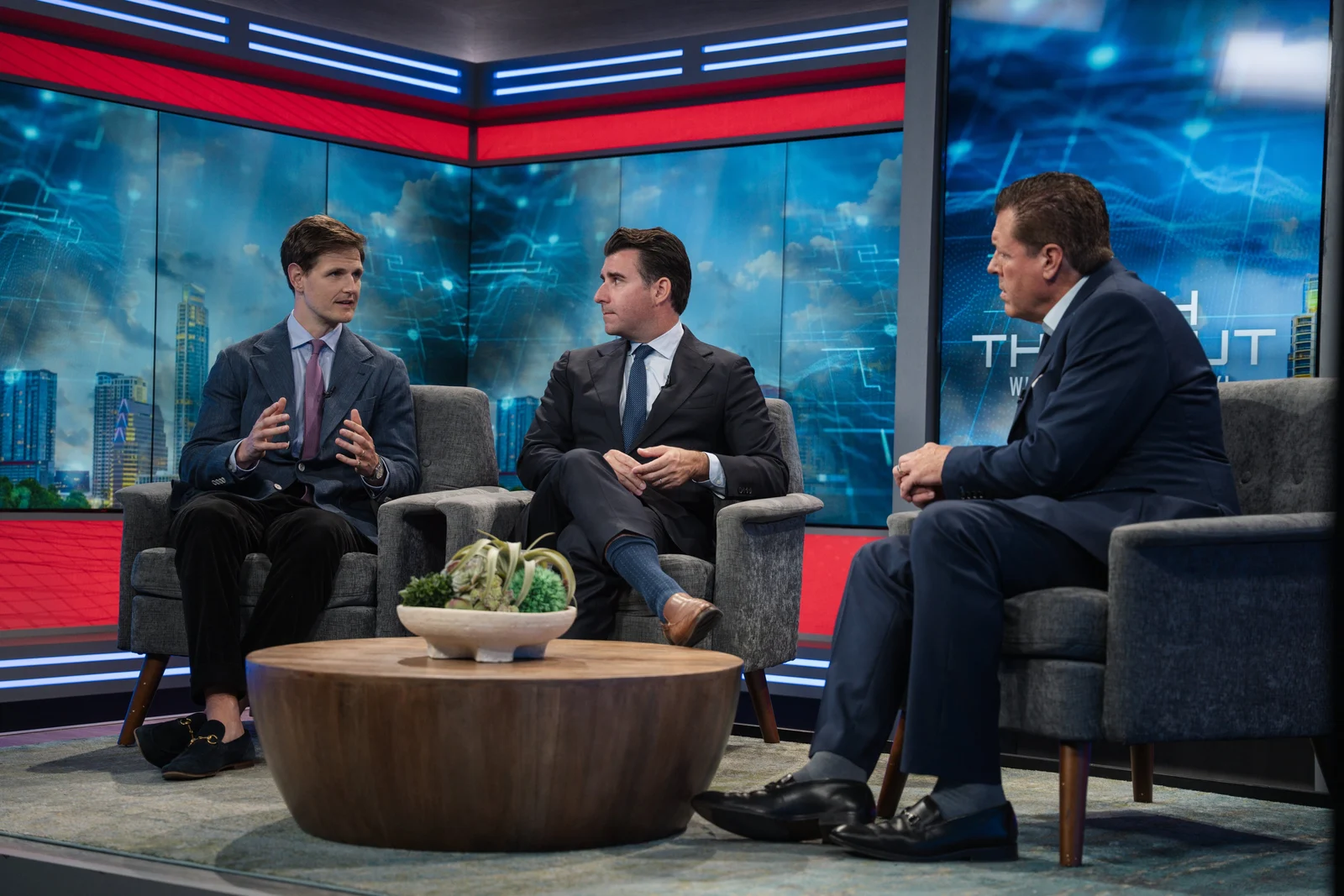 Three men seated in a television studio discussing the campus, an Austin skyline graphic on the screens behind them
